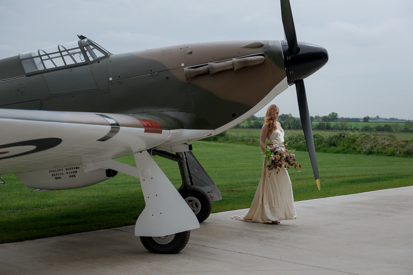 Into The Clouds - A 1940s Inspired Shoot | Ps With Love Jewellery and Accessories Bride in pale gold gown poses by the propellor of a Hurricane aircraft